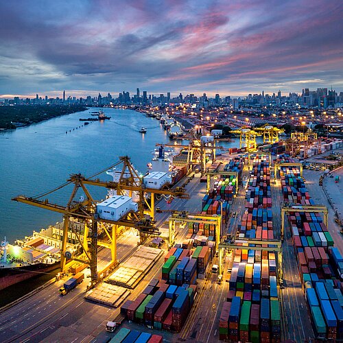 Aerial view of port with crane loading containers onto ship, against backdrop of Bangkok skyline at night.